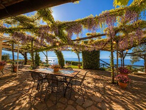 An outdoor patio with a table and chairs, surrounded by greenery and a view of the sea.