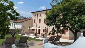A poolside area with lounge chairs and a tent, a building with red shutters, and a tree in the background. - Corte Massimago (Mezzane di Sotto)