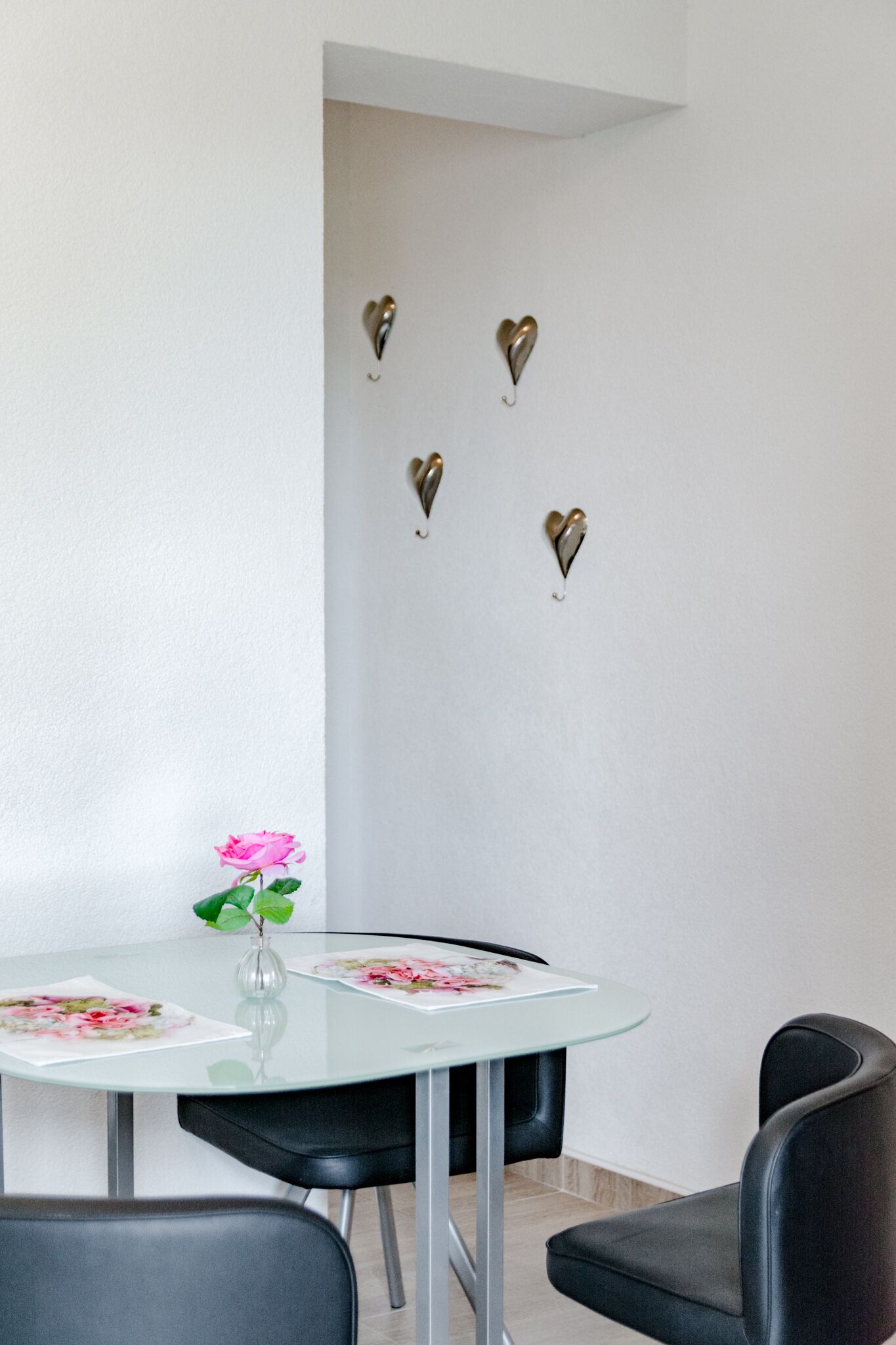 En matplats med ett glasbord, svarta stolar, en vas med en rosa blomma och hjärtformade väggdekorationer.