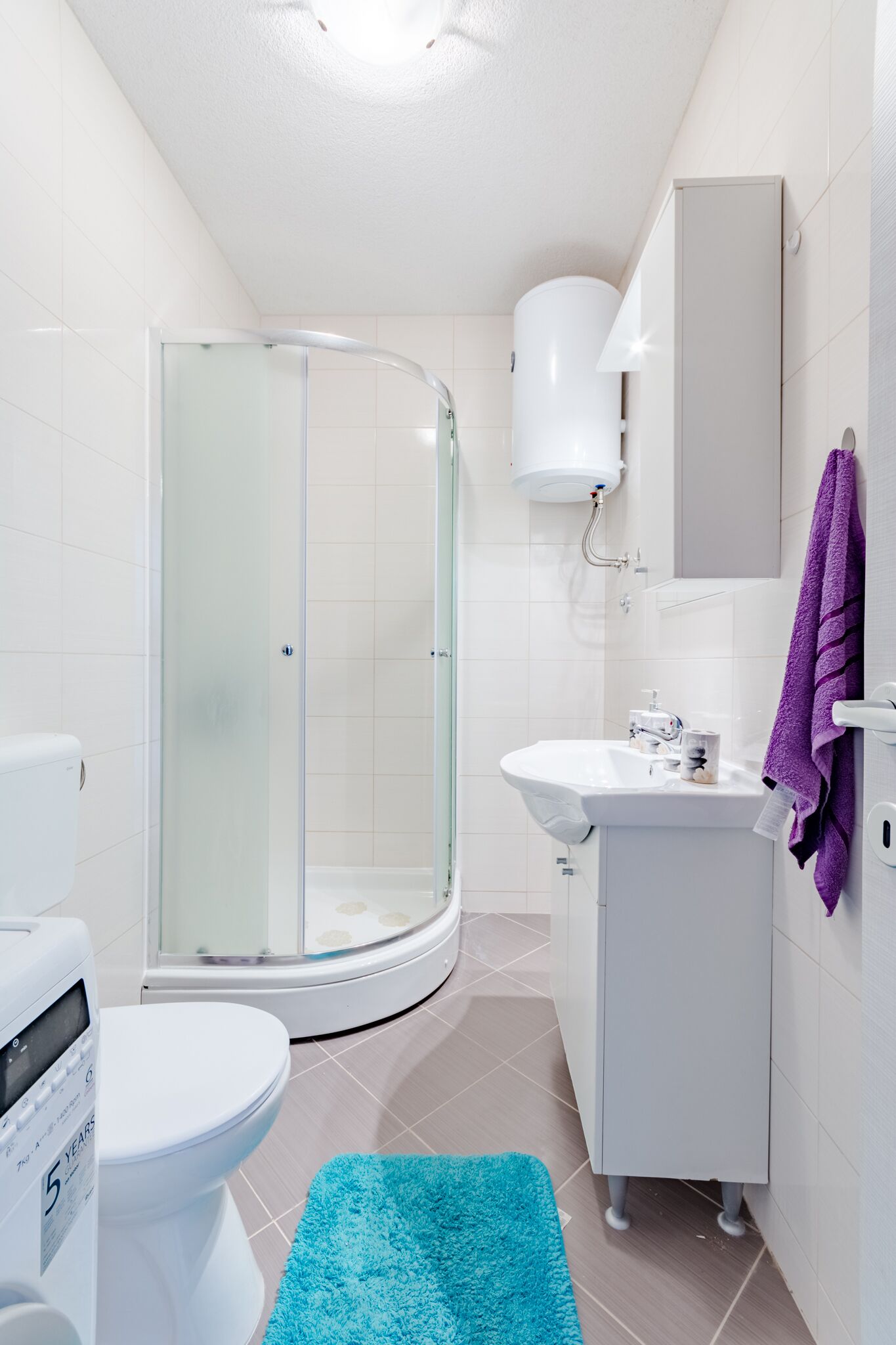 Ein modernes Badezimmer mit Duschkabine aus Glas, einer weißen Toilette, einem Waschbecken mit Spiegel und einem violetten Handtuch an der Wand.