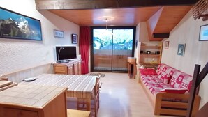 A cozy living room with a wooden dining table, a sofa with heart-patterned cushions, a TV, and a balcony with a red curtain. - South expo studio, heart resort, calm, ski to door (Les Deux Alpes)