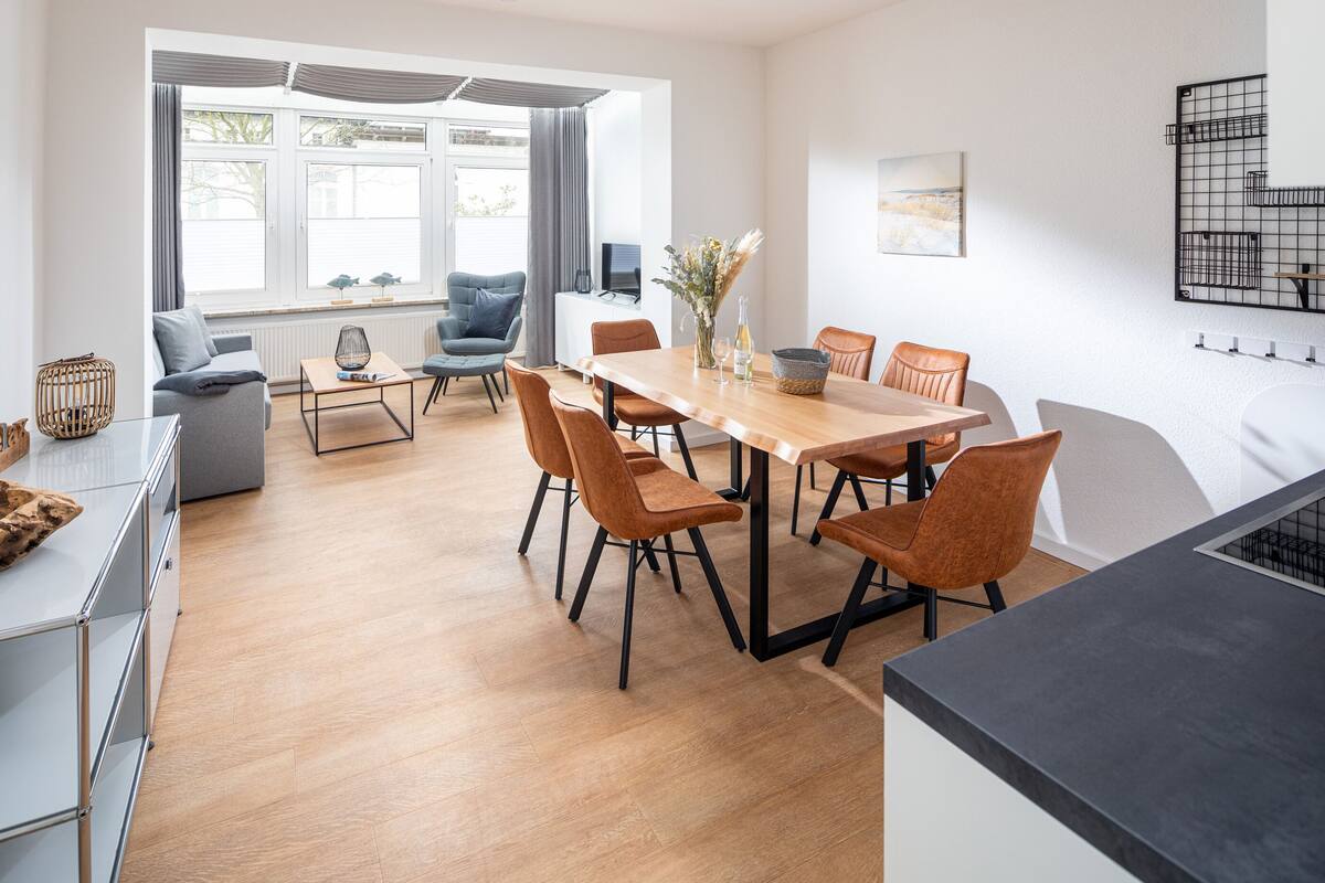 A modern dining area with a wooden table and orange chairs, a grey sofa, and a window with curtains.