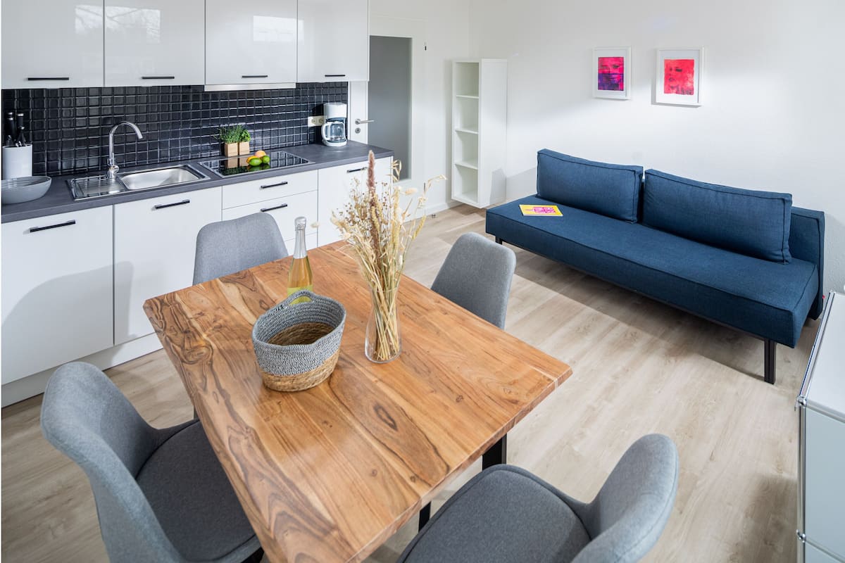 A modern kitchen with white cabinets, a black backsplash, and a dining area with a wooden table and grey chairs. There's a blue sofa and a white cabinet in the living area.