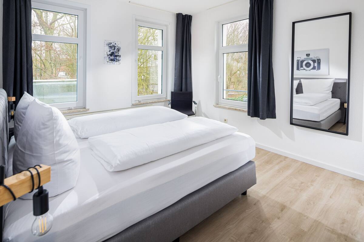 A modern bedroom with a large bed, two windows, a chair, and a mirror reflecting the bed.