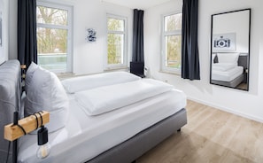 A modern bedroom with a large bed, two windows, a chair, and a mirror reflecting the bed. - Strandloft Drei Norderney (Norderney)