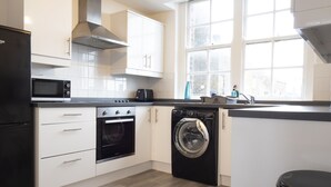 Eine moderne Küche mit weißen Schränken, einem schwarzen Kühlschrank, einer Mikrowelle, einem Backofen, einer Waschmaschine und einem Fenster mit Tageslicht.