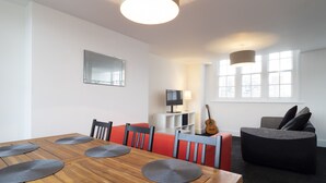 Ein modernes Wohnzimmer mit einem Holzesstisch, roten und schwarzen Stühlen, einem grauen Sofa, einer Gitarre und einem Fernseher.