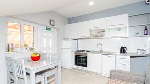 A compact kitchen with white cabinets, a dining table with chairs, a red fruit bowl, and a built-in oven. - Apartments Donita (Dubrovacko Primorje)