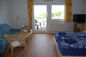 Una habitación con cama, sofá, mesa y sillas. Cuenta con una ventana con cortinas, un radiador y un televisor sobre un mueble.