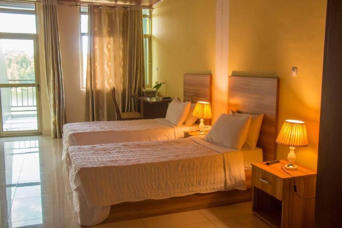 Habitació d'hotel amb dos llits, una tauleta de nit, una làmpada i vista a l'exterior.