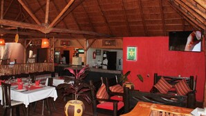 A restaurant with wooden tables and chairs, a thatched roof, and a TV mounted on the wall. - Delta Resort Hotel (Bwishyura)