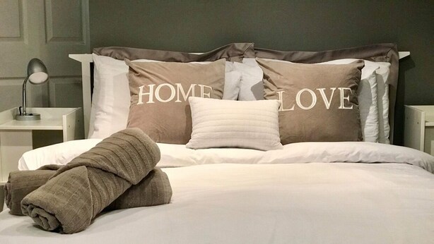 Un lit bien fait, avec du linge de lit blanc et une serviette marron, une lampe de chevet et des oreillers sur lesquels on peut lire les mots « HOME LOVE ».