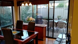 A dining area with a glass table and chairs, a vase of flowers, and a view of the backyard through large windows. - Modern Melville Plaza Townhouse close to Swan River and Port beach. (Palmyra)