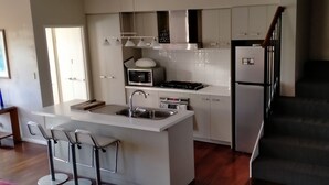 A modern kitchen with white cabinets, a built-in microwave, a stove, and a refrigerator. There is a small island with a sink and two bar stools. A staircase is visible to the right. - Modern Melville Plaza Townhouse close to Swan River and Port beach. (Palmyra)