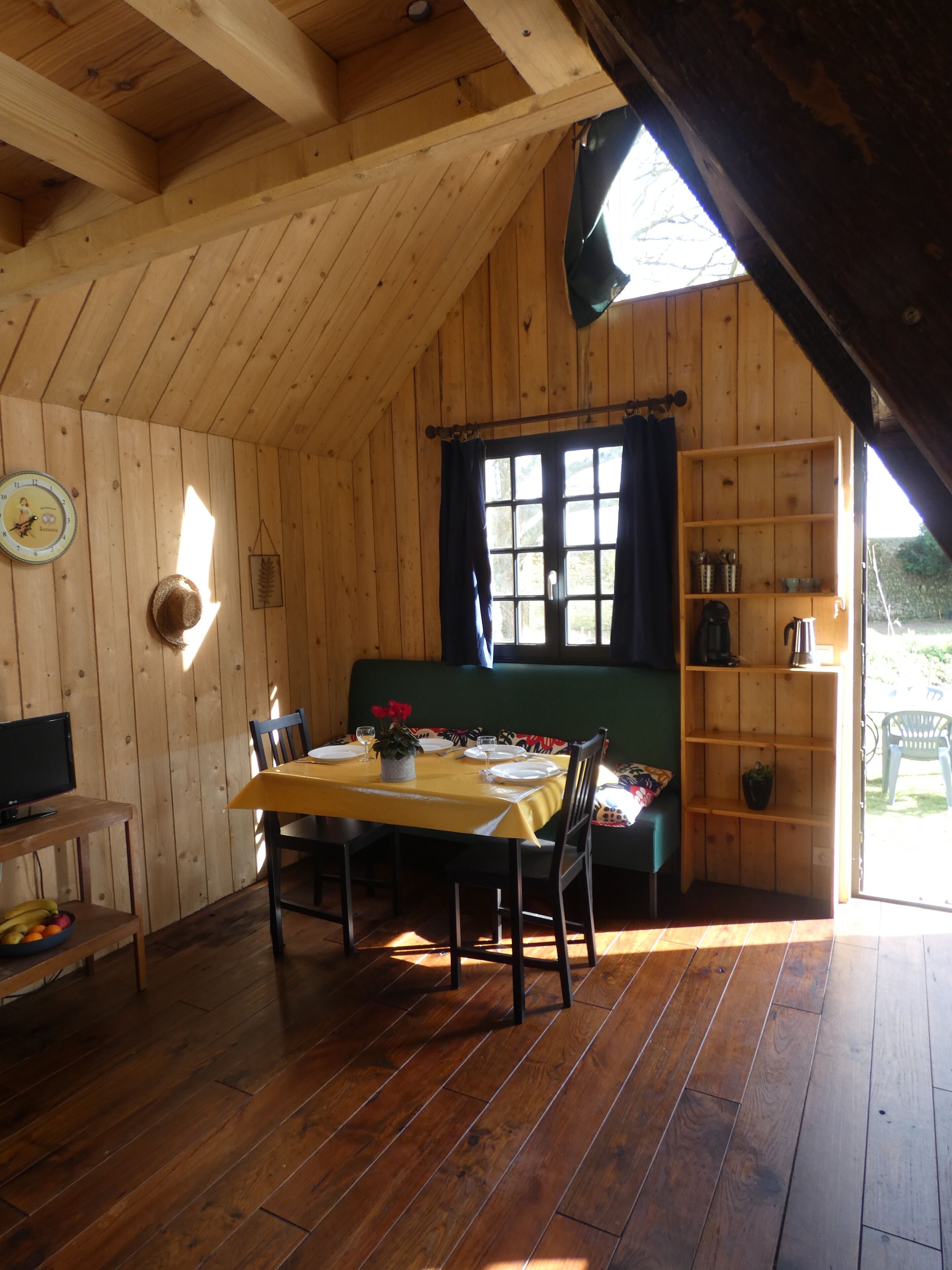 Une cabane en bois dotée d’un coin repas avec une table dressée pour deux personnes, d’un canapé vert et d’une étagère en bois où sont disposés divers objets.