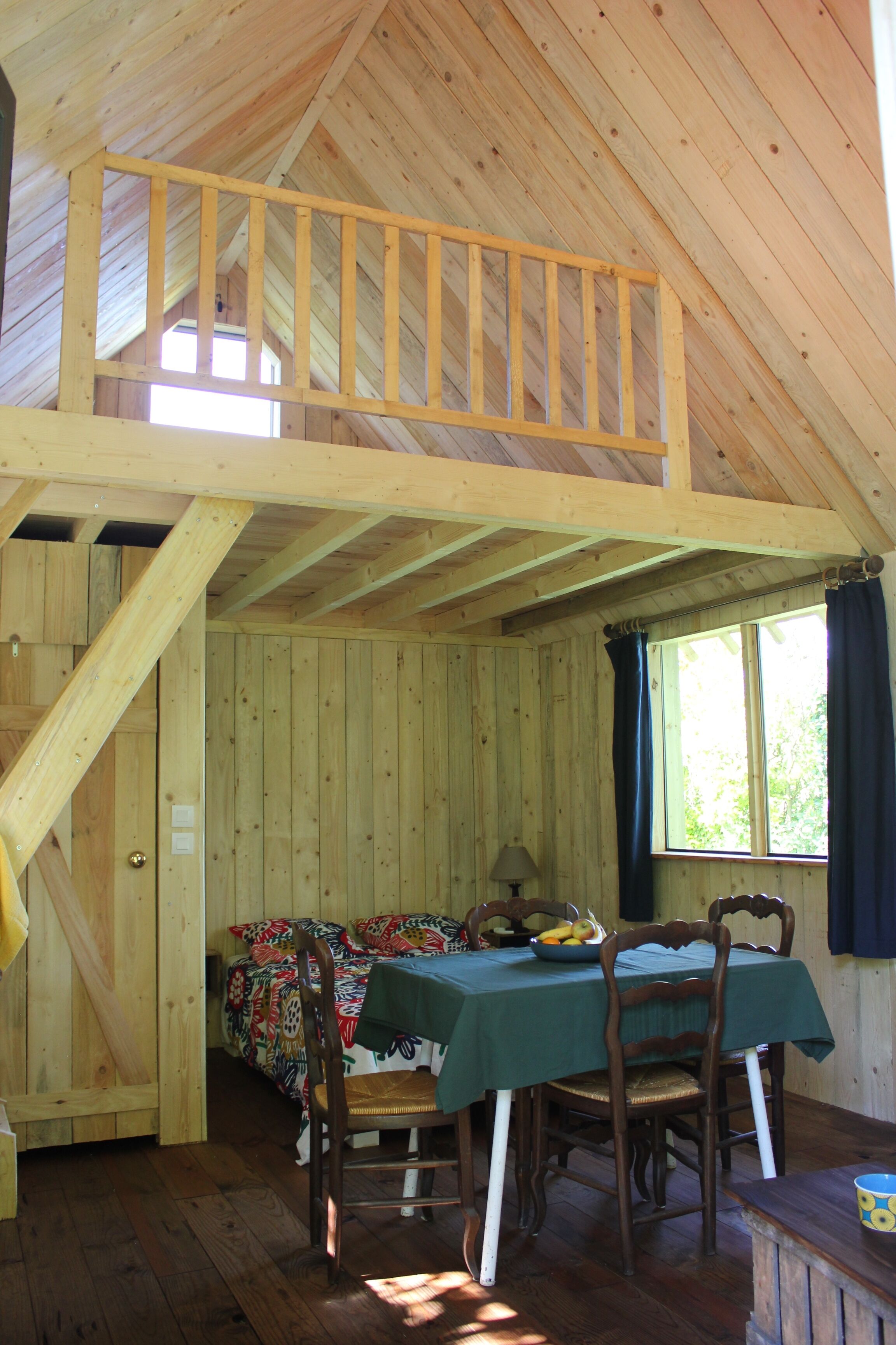 Une cabane en bois avec une chambre à coucher située en mezzanine, un coin repas avec une table et des chaises, et une fenêtre avec des rideaux.