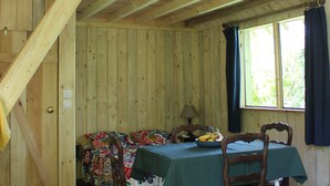 Een houten chalet met een slaapkamer op de zolder, een eethoek met tafel en stoelen, en een raam met gordijnen.