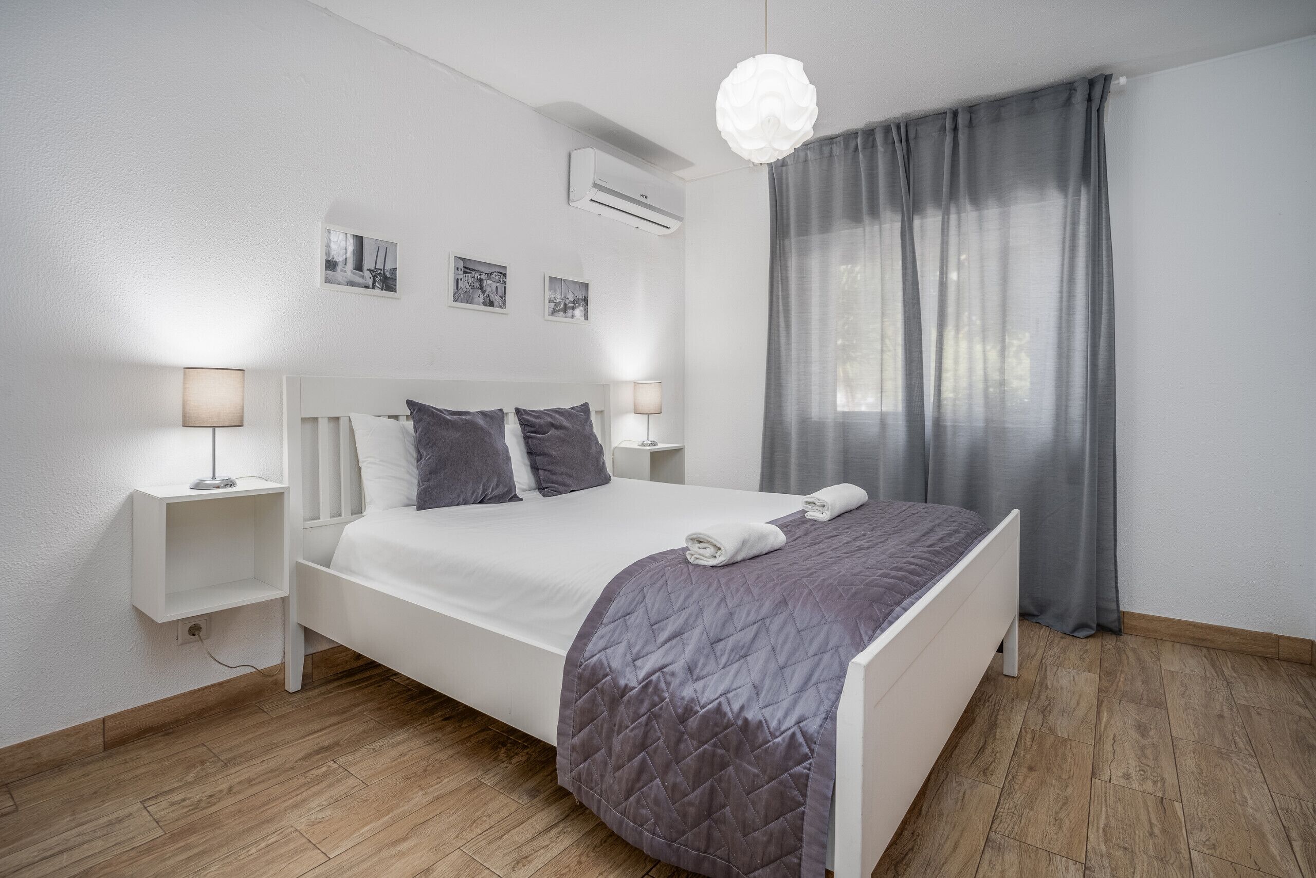 A modern bedroom with a queen-sized bed, white headboard, two bedside tables with lamps, a grey quilted bedspread, and a large window with grey curtains.