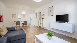 Un salon moderne avec un canapé gris, une table basse blanche agrémentée d’une plante, un téléviseur fixé au mur et un coin repas avec des chaises et une table.