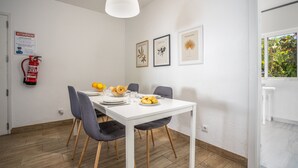 Un coin repas avec une table blanche, des chaises grises et un luminaire suspendu blanc. On peut voir des tableaux encadrés au mur, un extincteur et une fenêtre donnant sur un paysage verdoyant.