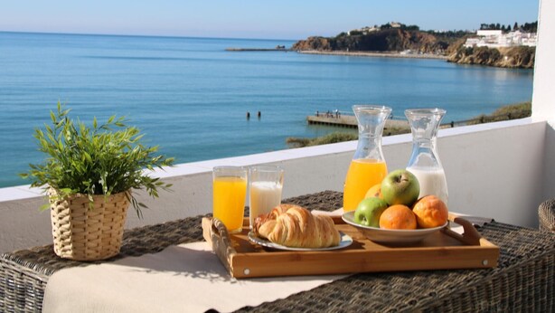 A breakfast tray with croissants, fruit, juice, and milk on a balcony overlooking a coastal view.