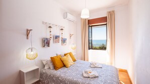 A bedroom with a bed, a bedside table, a hanging light fixture, and a window with a view of the sea.