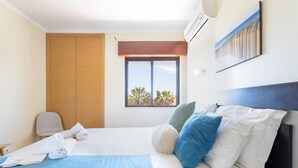 Une chambre d’hôtel avec un lit, une fenêtre donnant sur des palmiers, une armoire en bois et un climatiseur fixé au mur.