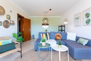 Una sala de estar con un sofá azul, una silla de mimbre, una mesita y una lámpara colgante.