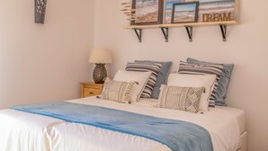 Ein ordentlich bezogenes Bett mit blauer Decke, ein Nachttisch mit Lampe und eine Wandregal mit gerahmten Bildern und einem 'DREAM' Schild.