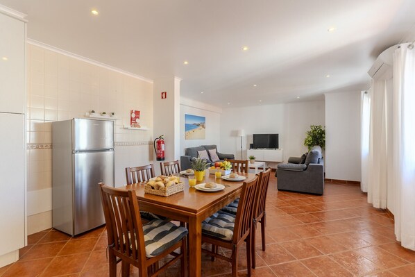 A modern kitchen with a refrigerator, dining table with chairs, and a living area with a sofa and TV.
