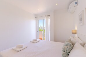 A hotel room with a bed, a window with curtains, a wall-mounted air conditioner, and a bedside lamp.