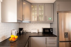 A kitchen with grey cabinets, a stainless steel refrigerator, a toaster, a coffee maker, and a bowl of fruit on the counter.