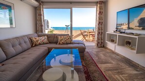 Een woonkamer met een bank, een glazen salontafel en een televisie aan de muur. Er is een groot raam met uitzicht op het strand.