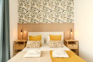 Une chambre d’hôtel avec un lit, deux tables de chevet, une tête de lit en bois et un papier peint à motifs de feuilles.