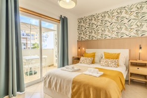Habitación de hotel con cama, mesita de noche de madera, una pequeña mesa y balcón con vistas a edificios.