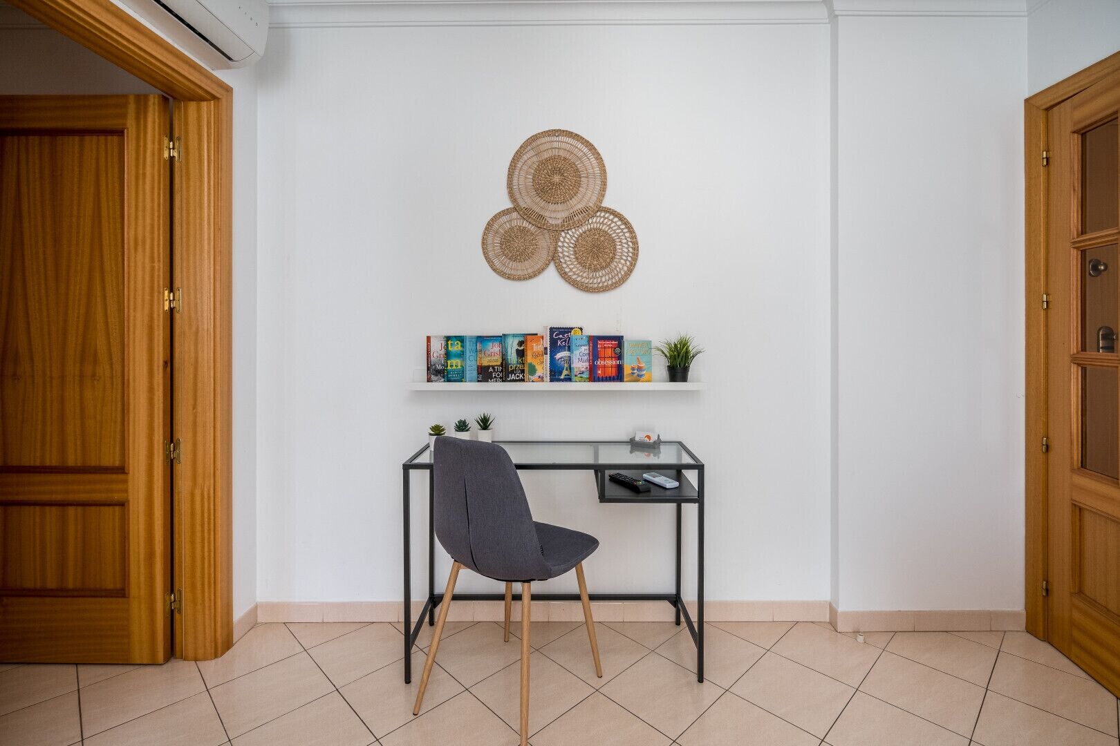 A room with a wooden door, a desk with a chair, a wall-mounted shelf with books and a small plant, and a decorative wall hanging.