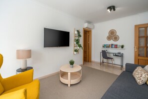 Un salon moderne avec un téléviseur à écran plat, une table basse ronde en bois, un fauteuil jaune, un canapé gris et un petit bureau avec une chaise.
