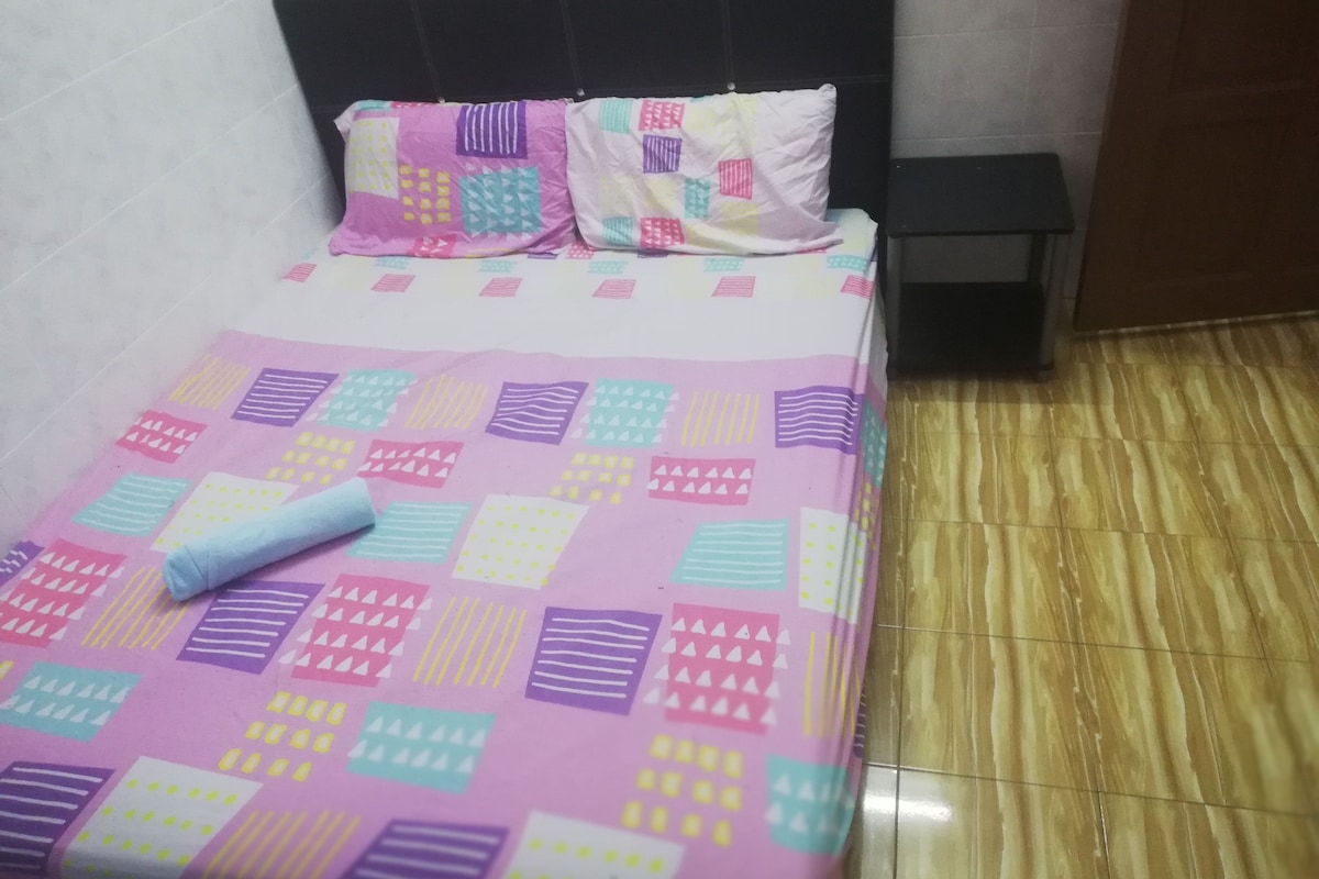 Una cama con una colcha y almohadas de estampado colorido, un cabecero negro, una mesita de noche pequeña y un suelo de baldosas.