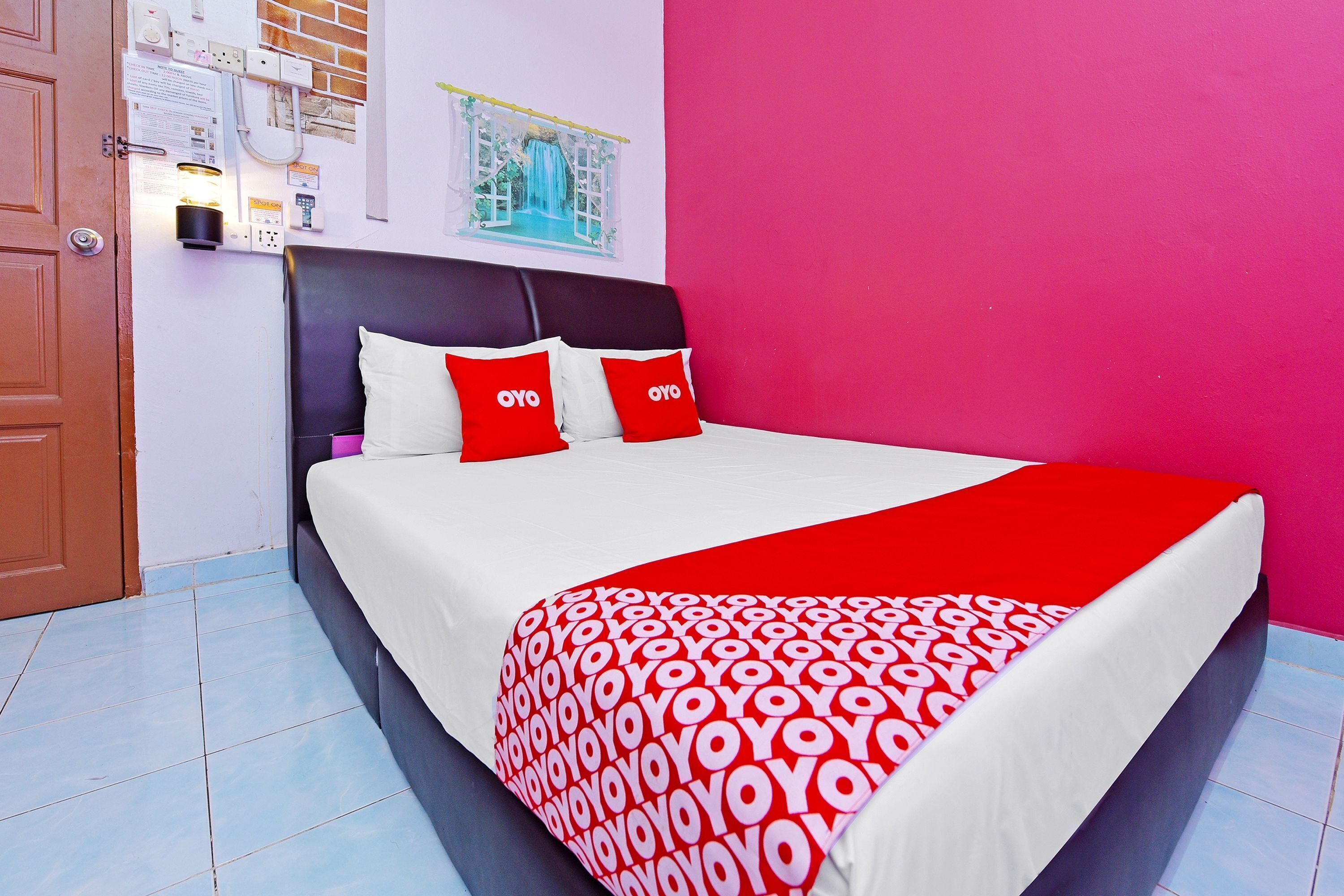 Una cama con una colcha de cuadros rojos y blancos, dos almohadas rojas con la marca 'OYO', una pared blanca con una ventana que muestra un cielo azul y una puerta a la izquierda.
