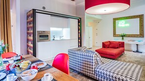 Een moderne woonkamer met een bank, een eettafel met ontbijt, een rode hanglamp en een spiegel met een neonbord.
