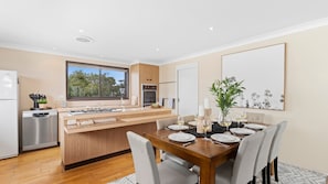 A modern kitchen with a central island, a dining area with a wooden table set for four, and a large window providing a view of the outdoors. - Watersedge  – absolute waterfront (Currarong)