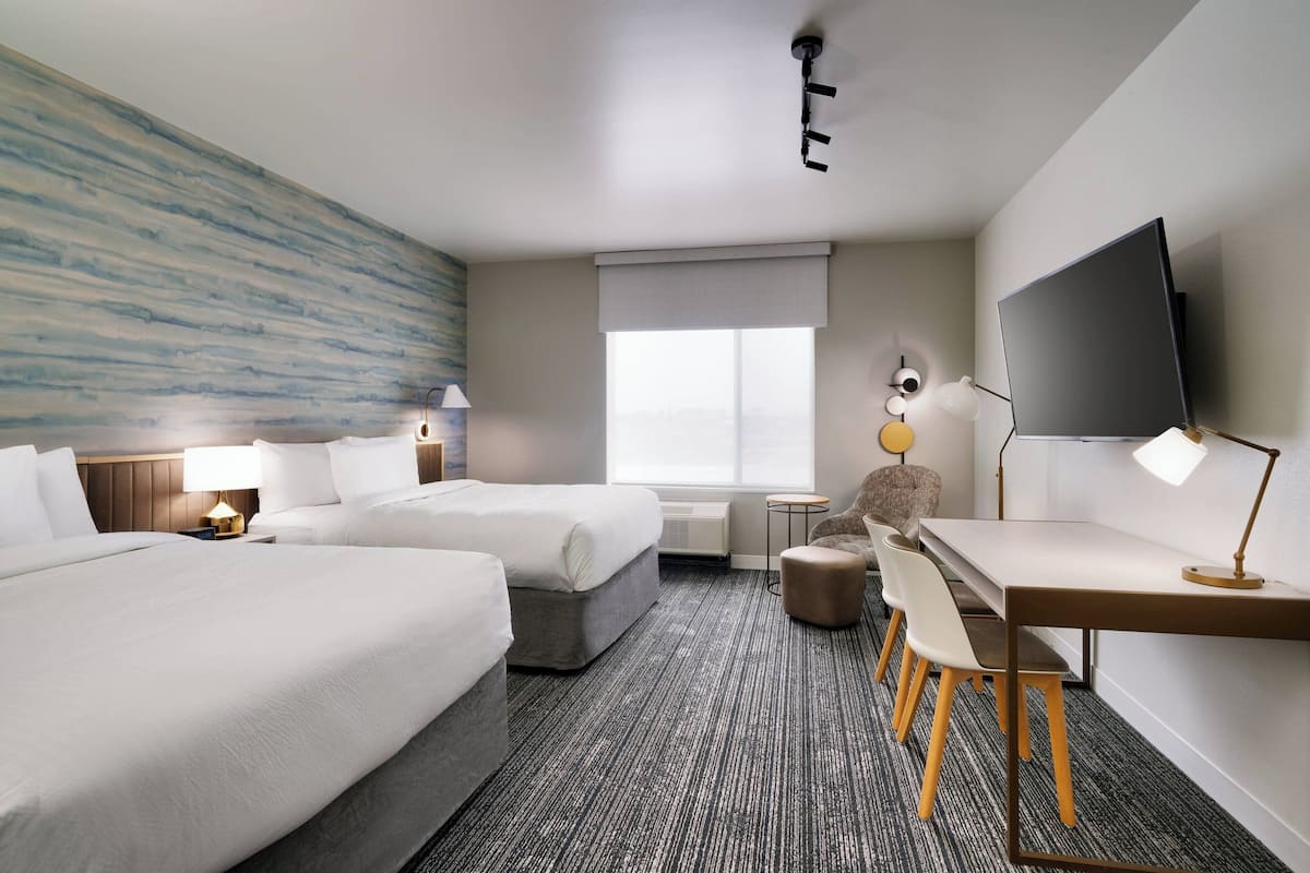 Ein Hotelzimmer mit zwei Betten, einem Schreibtisch, einem Sessel, einem Fernseher und einem Fenster.
