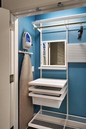 A bathroom with a blue wall, a mirror, an ironing board, and a shelf with storage bins. - TownePlace Suites by Marriott Salt Lake City Murray (Murray)