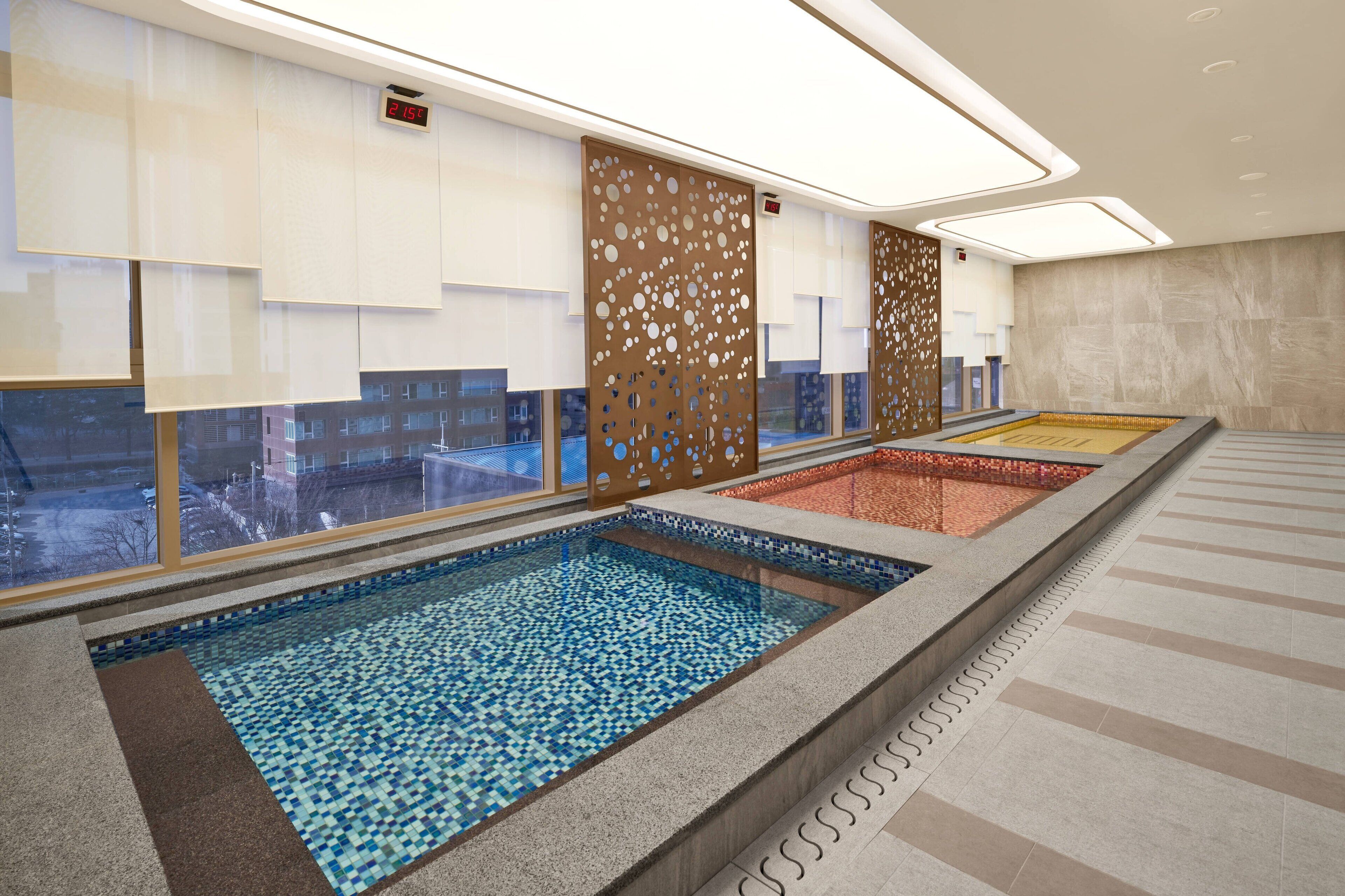 Una piscina interior moderna con suelo de baldosas, vista panorámica de la ciudad a través de amplios ventanales y un separador decorativo con círculos recortados.