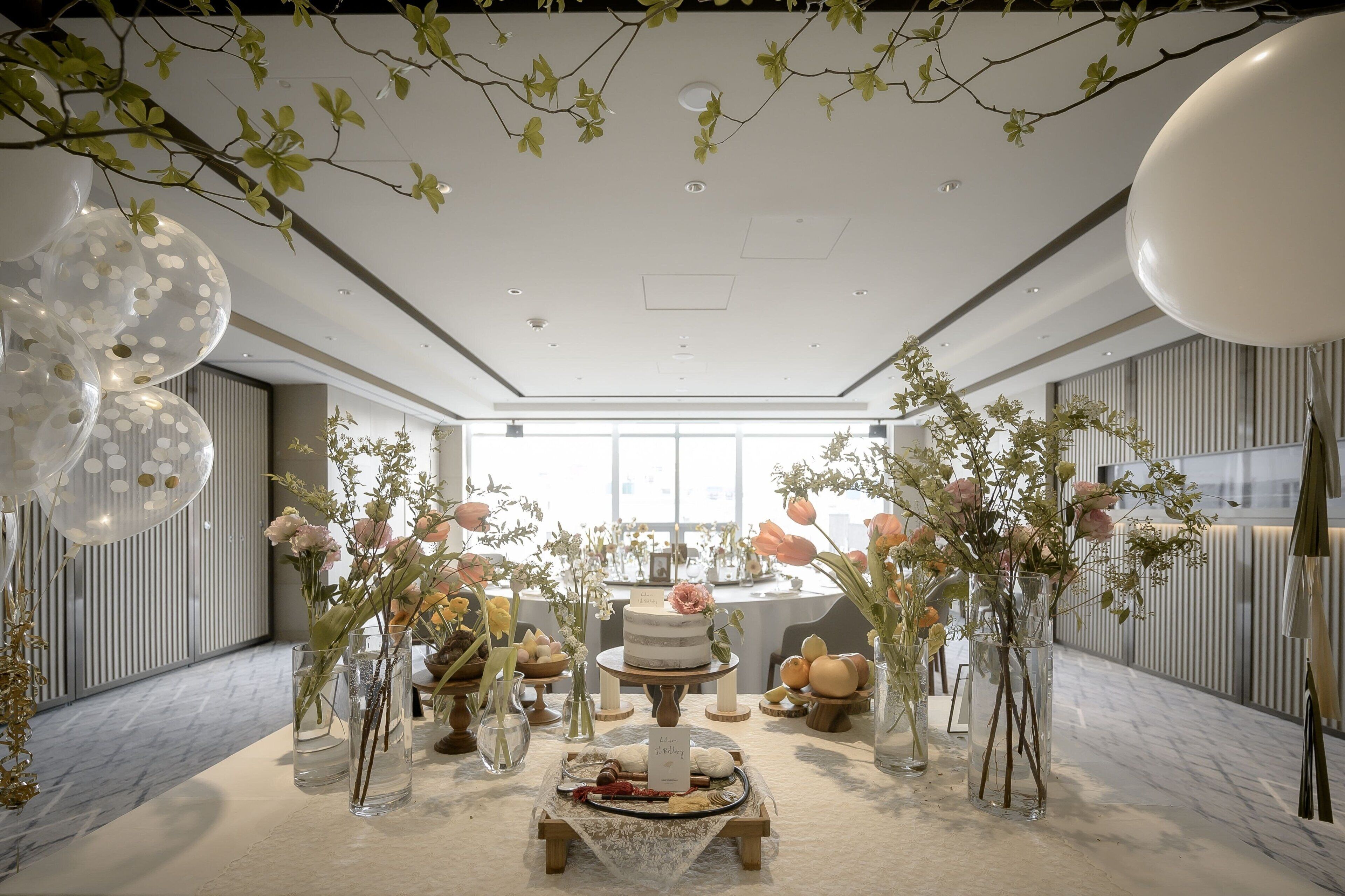 A modern, elegantly decorated indoor space with a central table featuring a cake, surrounded by floral arrangements and balloons.