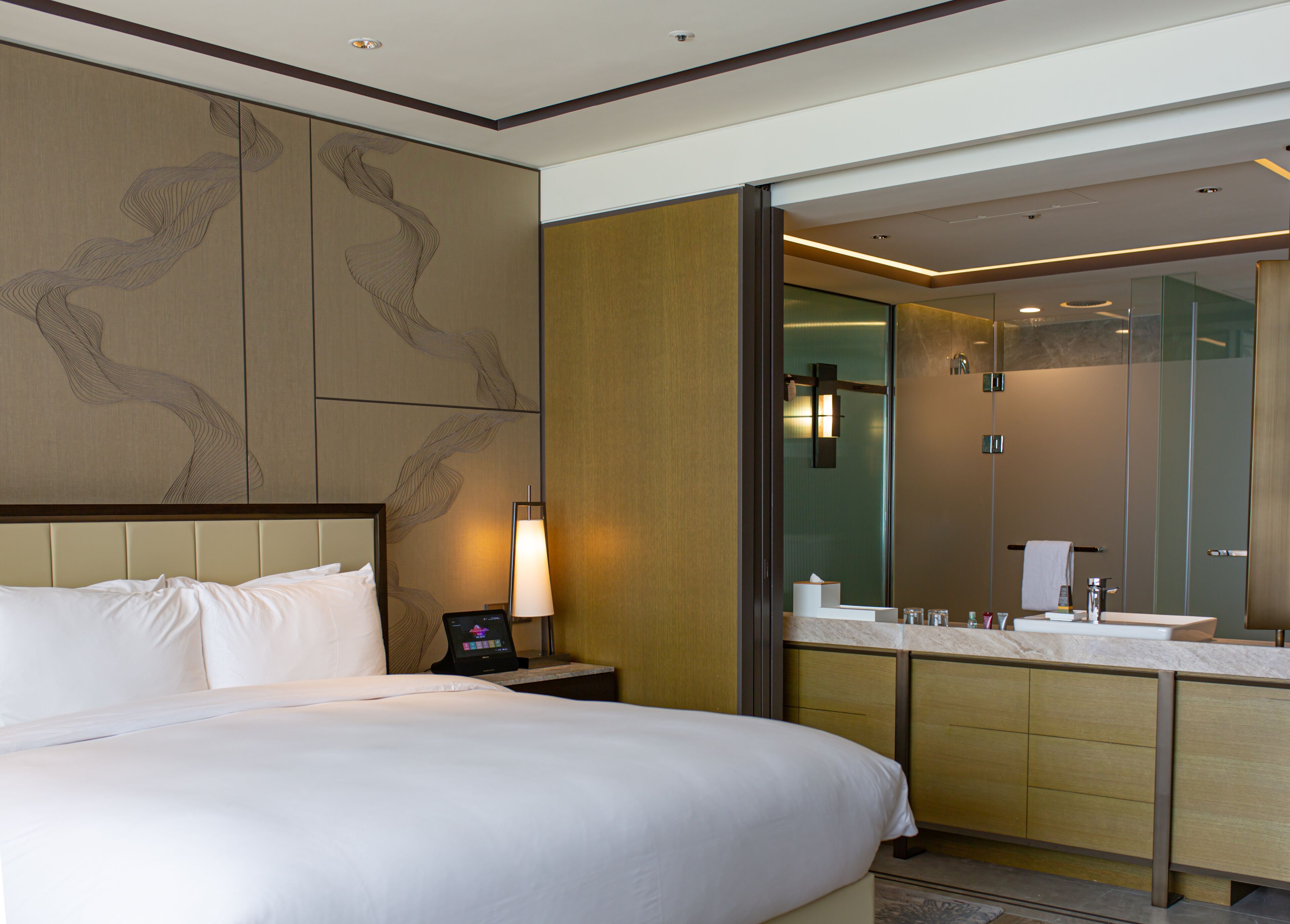A hotel room with a large bed, a bedside table with a lamp, a modern bathroom with a glass shower enclosure, and a large mirror.
