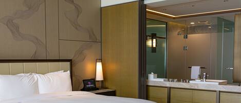 A hotel room with a large bed, a bedside table with a lamp, a modern bathroom with a glass shower enclosure, and a large mirror.