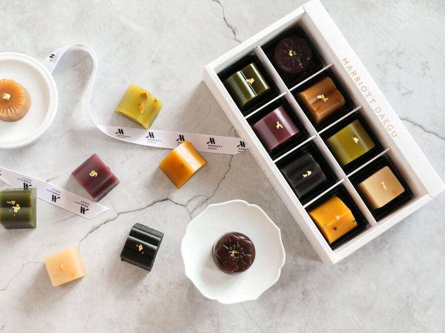 Una colección de velas de cera de distintas formas y colores, con una caja blanca que indica "MARRIOTT DAEGU" al fondo.