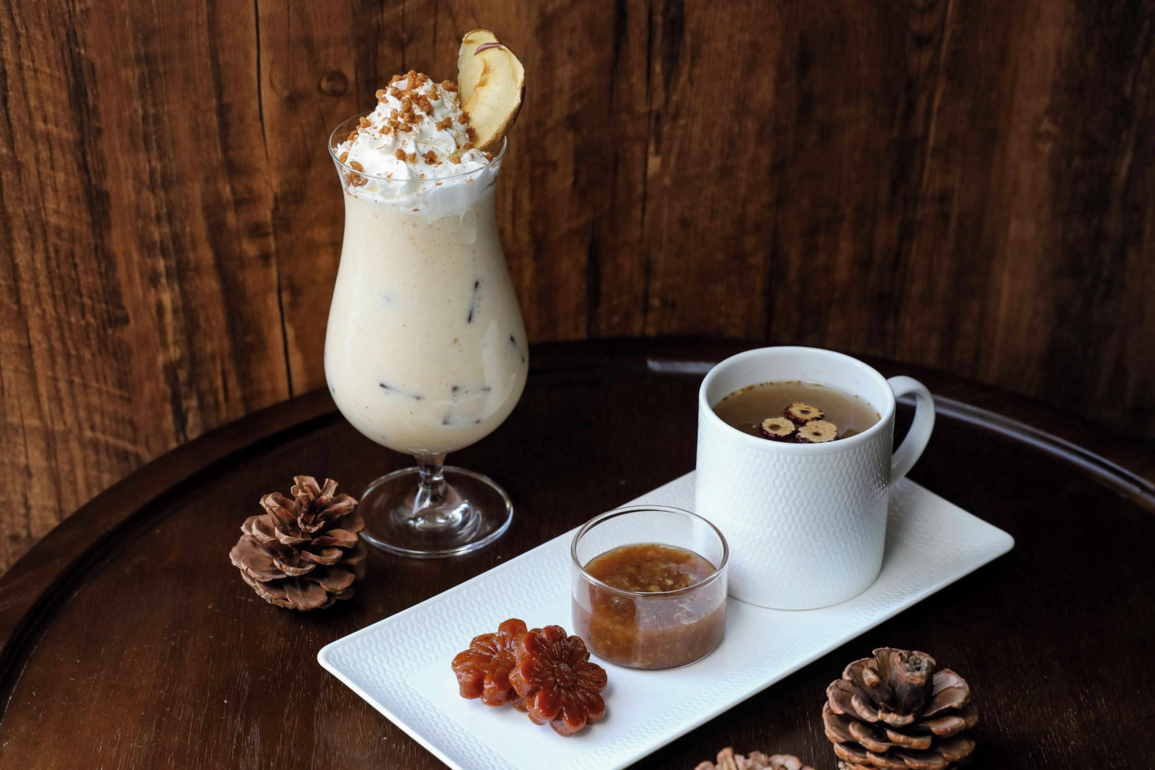Ein Dessertgetränk im Glas, eine Tasse mit einem Getränk und ein Teller mit einem kleinen Krug und Tannenzapfen auf einem Holztisch.