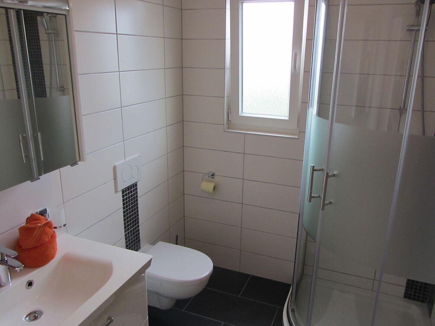 Baño con lavamanos blanco, inodoro, cabina de ducha con mampara de vidrio y ventana que proporciona luz natural.